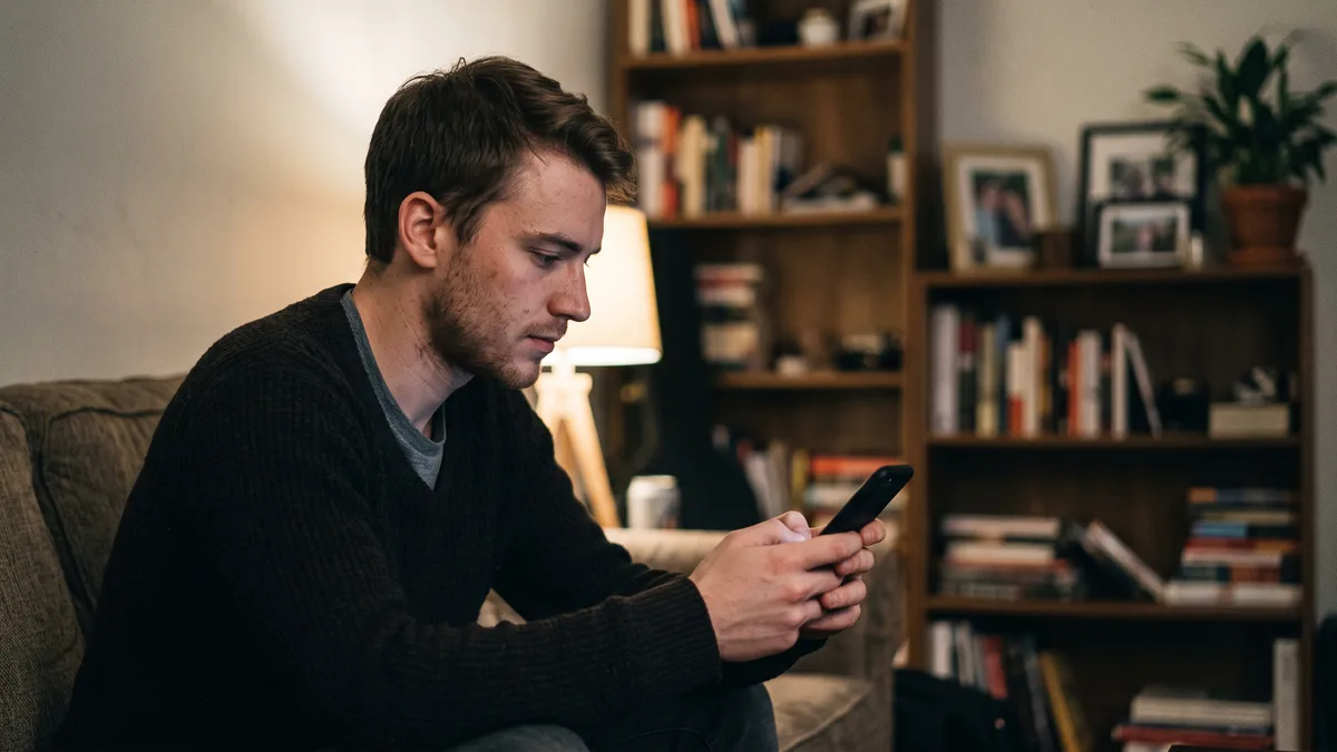 Man sitting on a sofa leaning forward, holding his phone in both hands while thinking about how to apologize